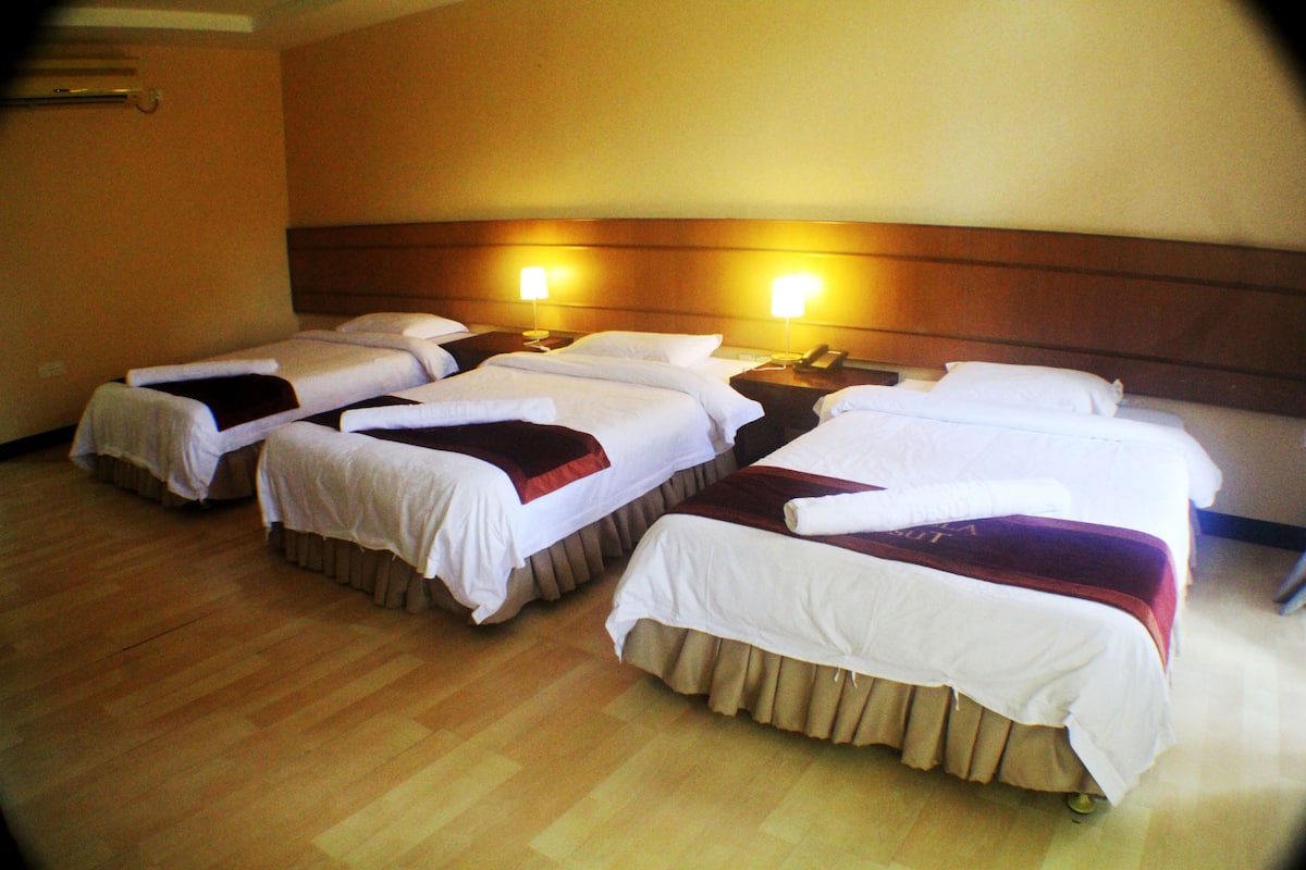 A hotel room with four single beds arranged in a row, each with a bedside table and lamp.