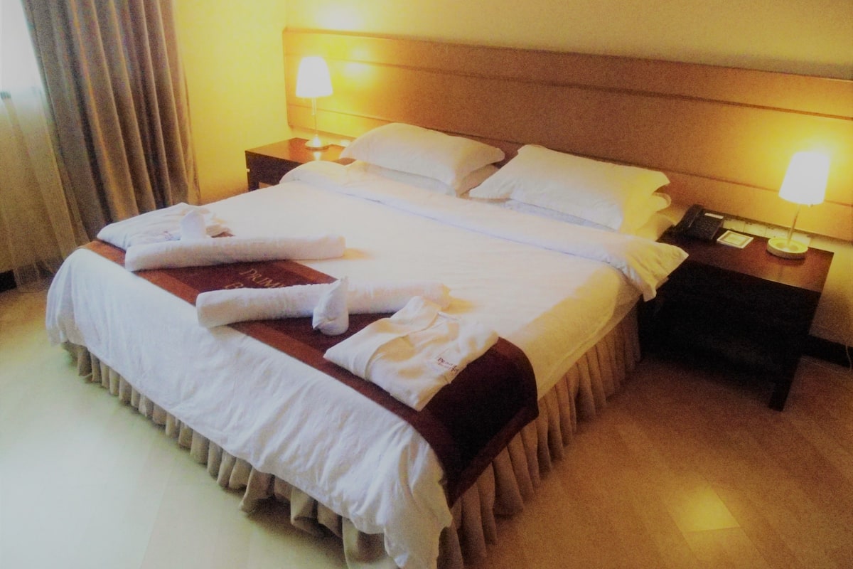 A neatly made bed with white linens and pillows, two bedside lamps, a bedside phone, and a small table with a lamp.