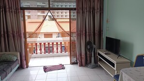 Una habitación con cama, televisión, ventilador y vistas a edificios a través de una ventana con cortinas.