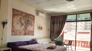 Habitación de hotel con un gran mapa mundial en la pared, una cama con ropa de cama morada y blanca, una mesita con una silla, y un balcón con vistas a edificios.