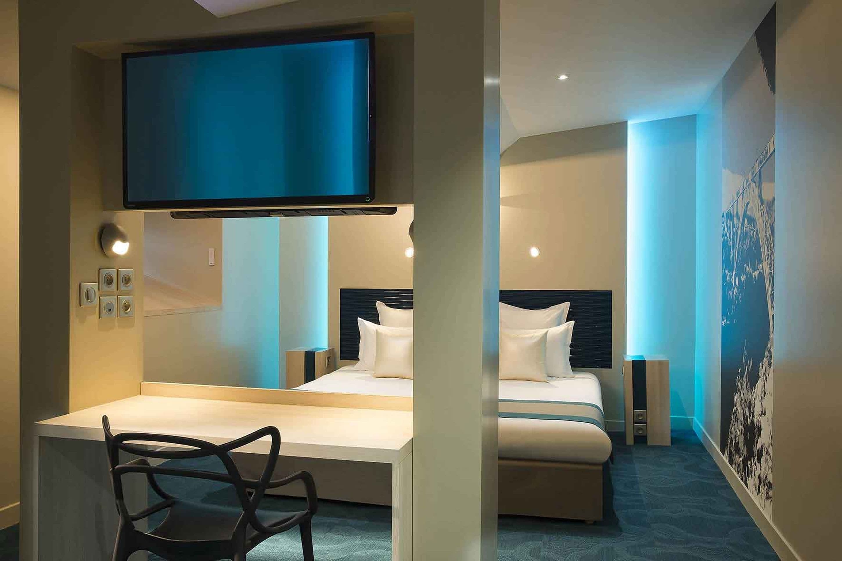 A modern hotel room with a large bed, a desk, and a flat-screen TV mounted above the bed.