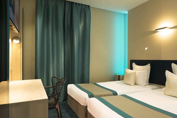 Ein Hotelzimmer mit zwei Betten, einem Schreibtisch, einem Stuhl und einem großen Fenster mit Vorhängen.
