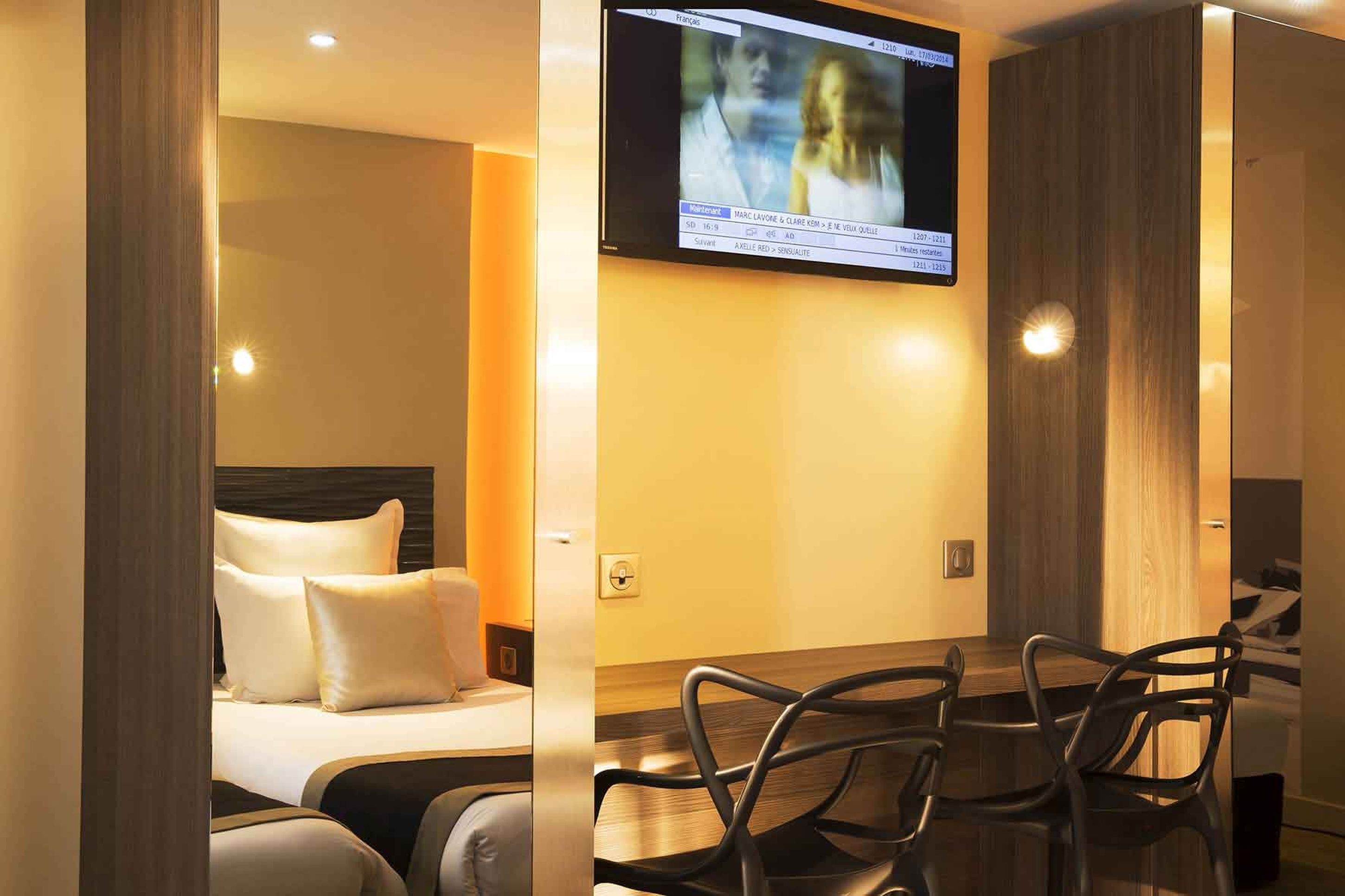 A hotel room with a bed, a TV mounted on the wall, and a dining area with chairs.