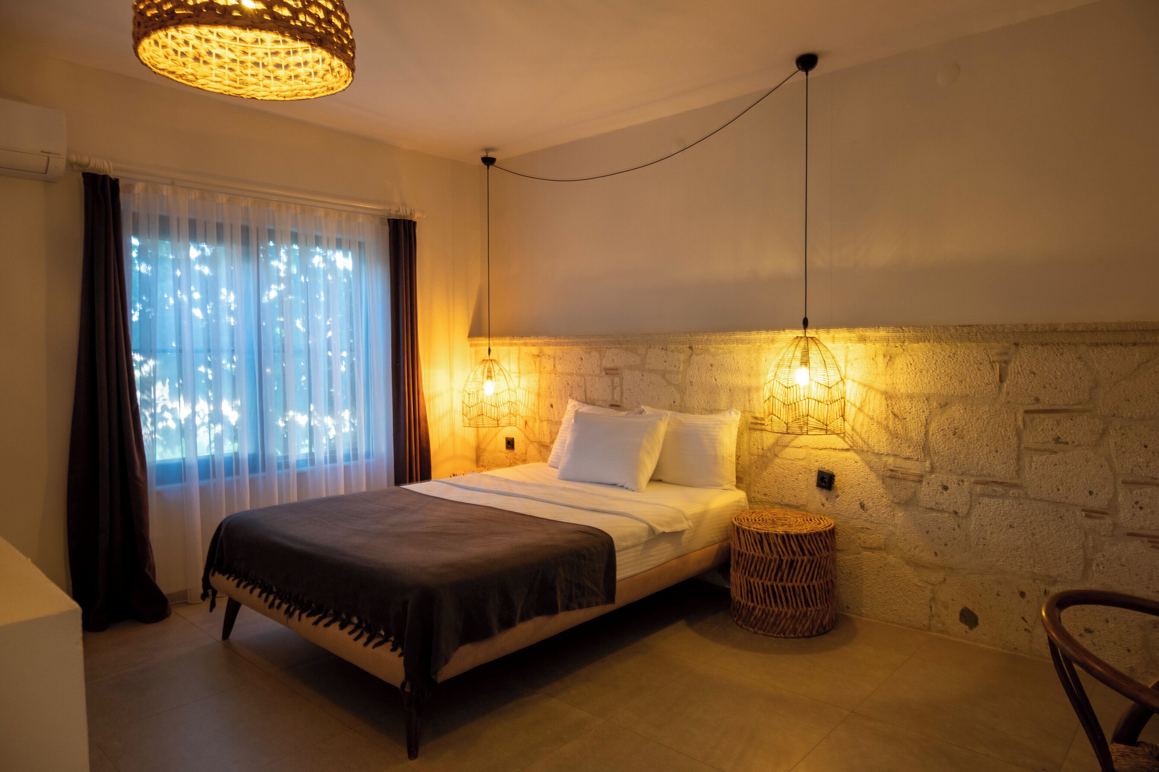 A bedroom with a bed, a wall-mounted lamp, a window with curtains, and a stone wall.