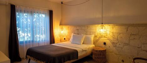 A bedroom with a bed, a wall-mounted lamp, a window with curtains, and a stone wall.