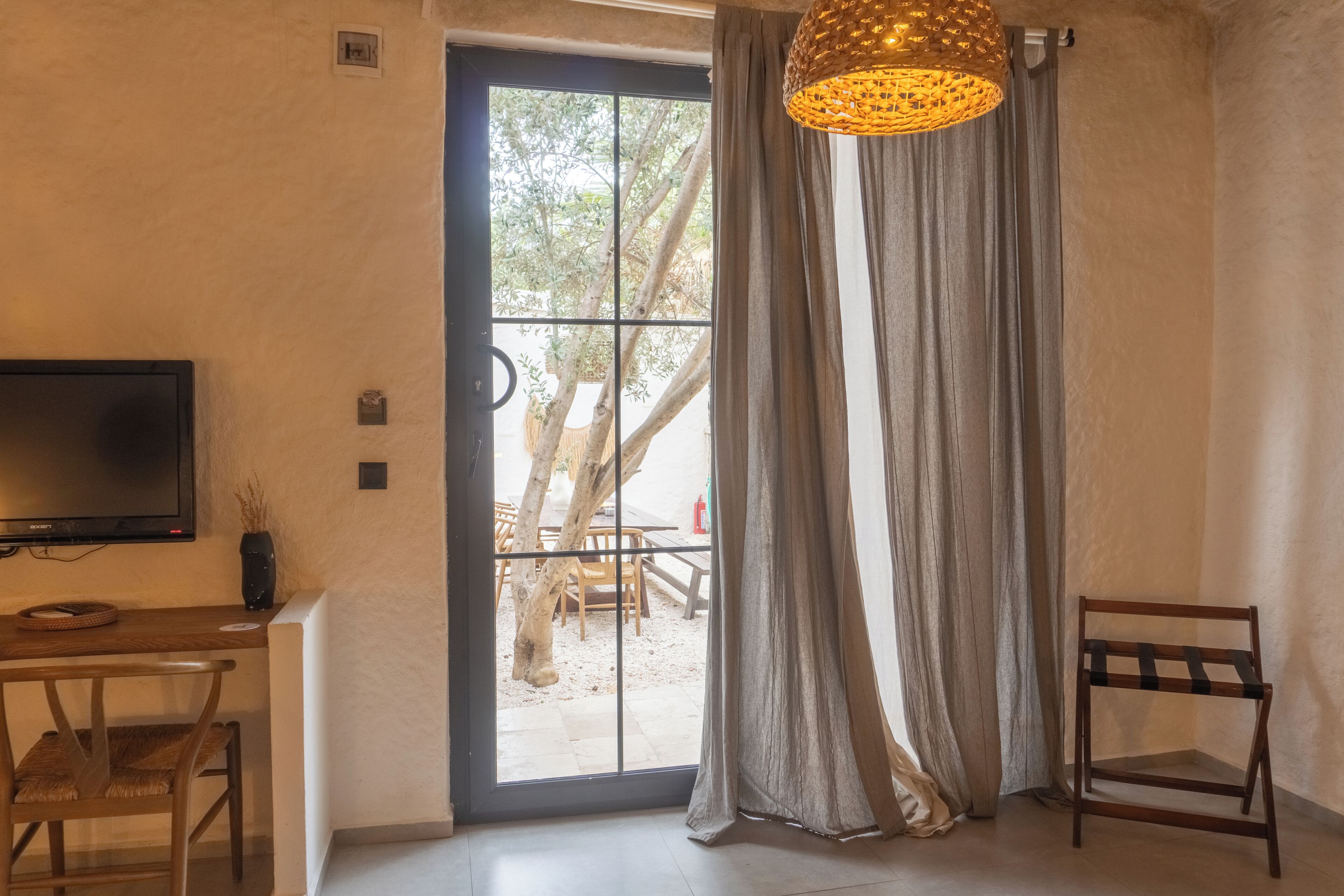 Una habitación con una televisión sobre un mueble bajo, una silla de madera, una mesa de comedor con sillas, un ventanal con cortinas y una lámpara colgante.