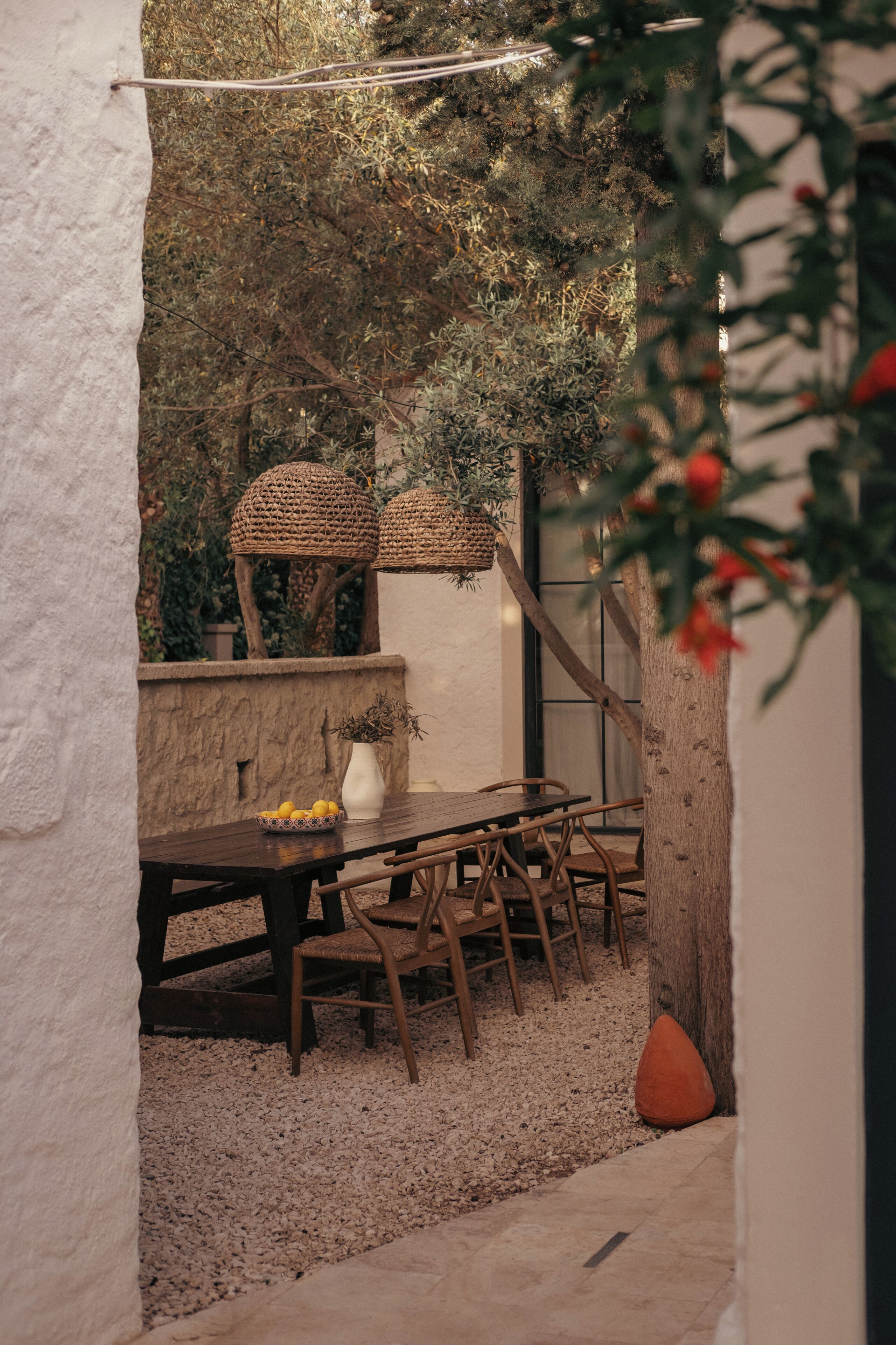 An outdoor dining area with a rustic wooden table and chairs, surrounded by a gravel floor and a white wall.