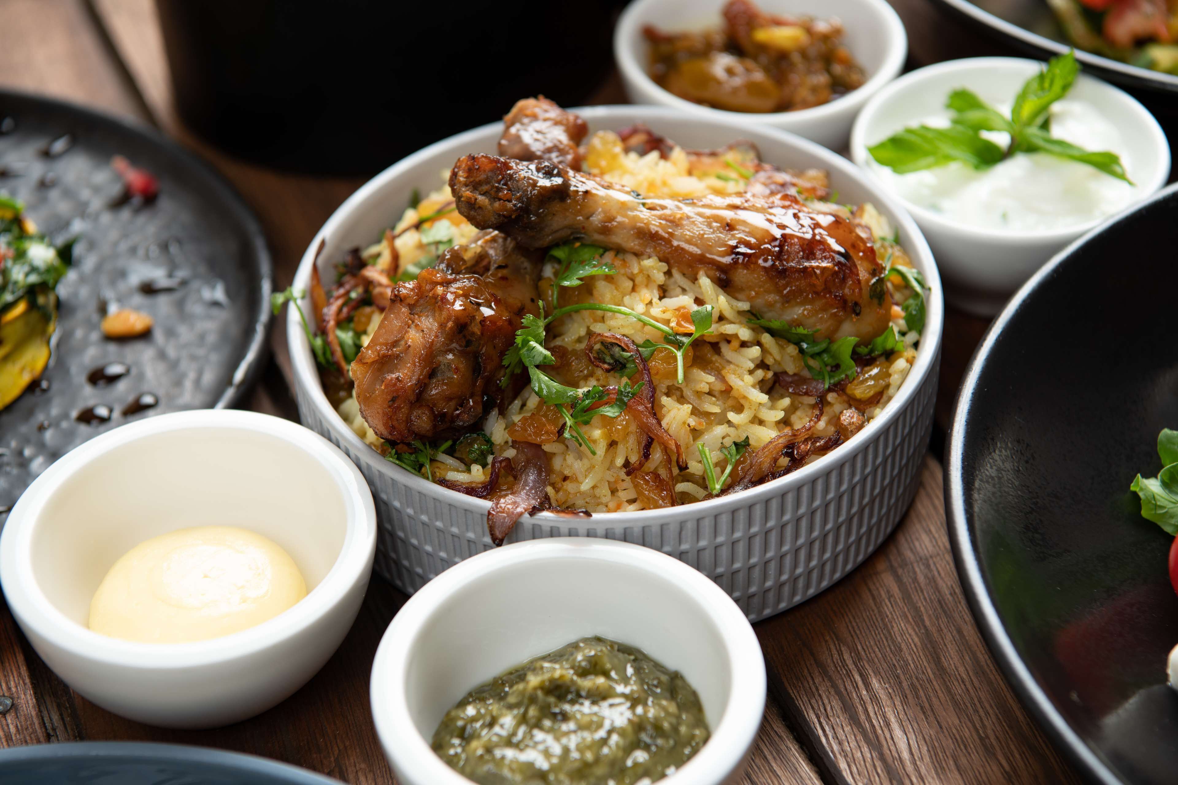 Un plato de biryani con pollo, adornado con hierbas, acompañado de pequeñas porciones de yogur y chutney.