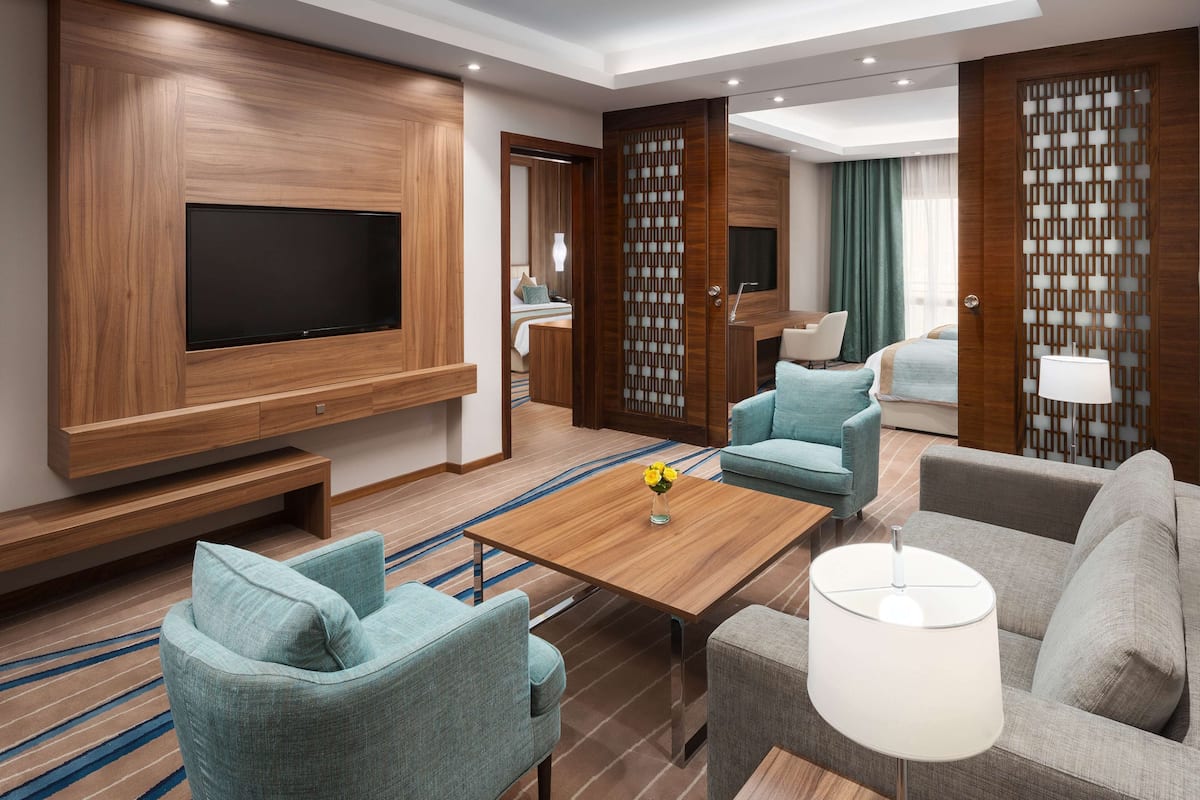 A modern hotel room with a large flat-screen TV mounted on a wooden wall, a wooden coffee table, two sofas, a small table with a vase of flowers, and a large window with curtains.