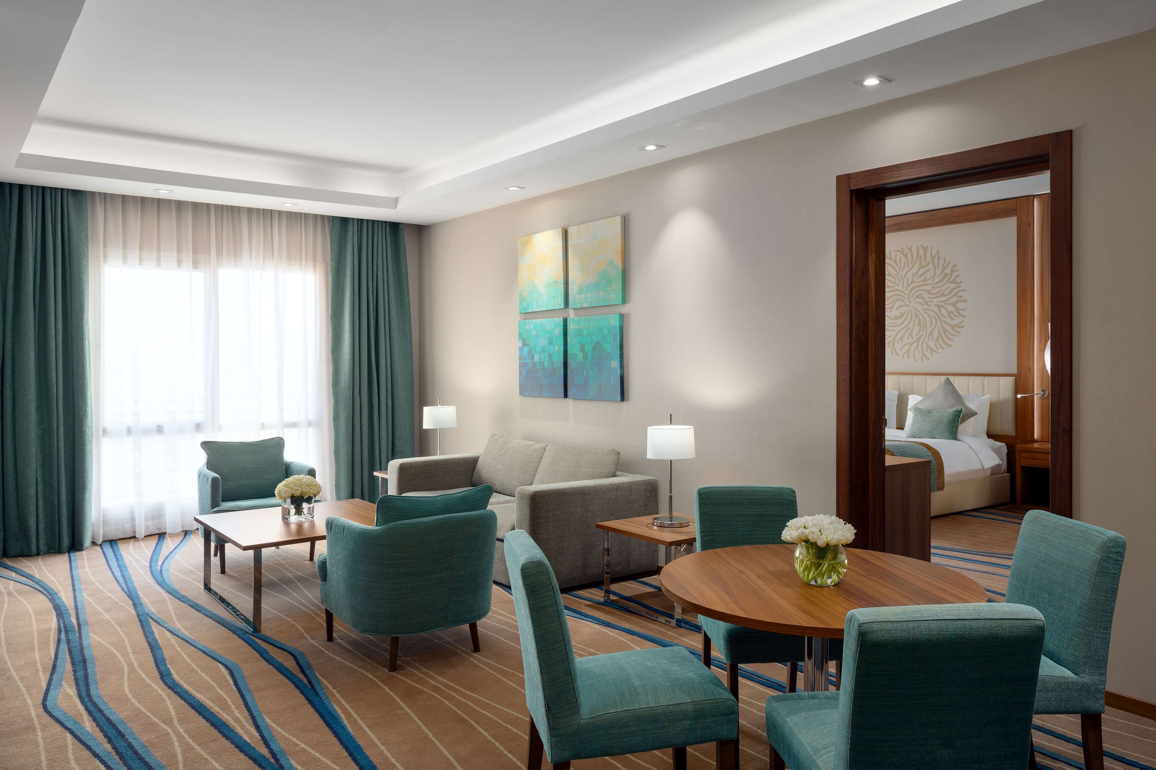 A hotel room with a sofa, two armchairs, a coffee table, and a dining table. There is a bed with a headboard and a nightstand. The room has a large window with curtains, a painting on the wall, and a decorative wall piece.