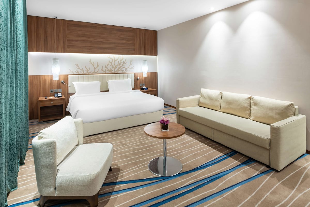 A modern hotel room with a large bed, a sofa, a chair, a small table, and a decorative wall panel.