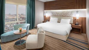 Hotelkamer met een groot bed, een bank, een stoel, een kleine tafel en uitzicht op de stad door grote ramen.