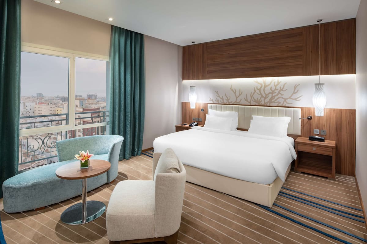 A hotel room with a large bed, a sofa, a chair, a small table, and a view of the city through large windows.