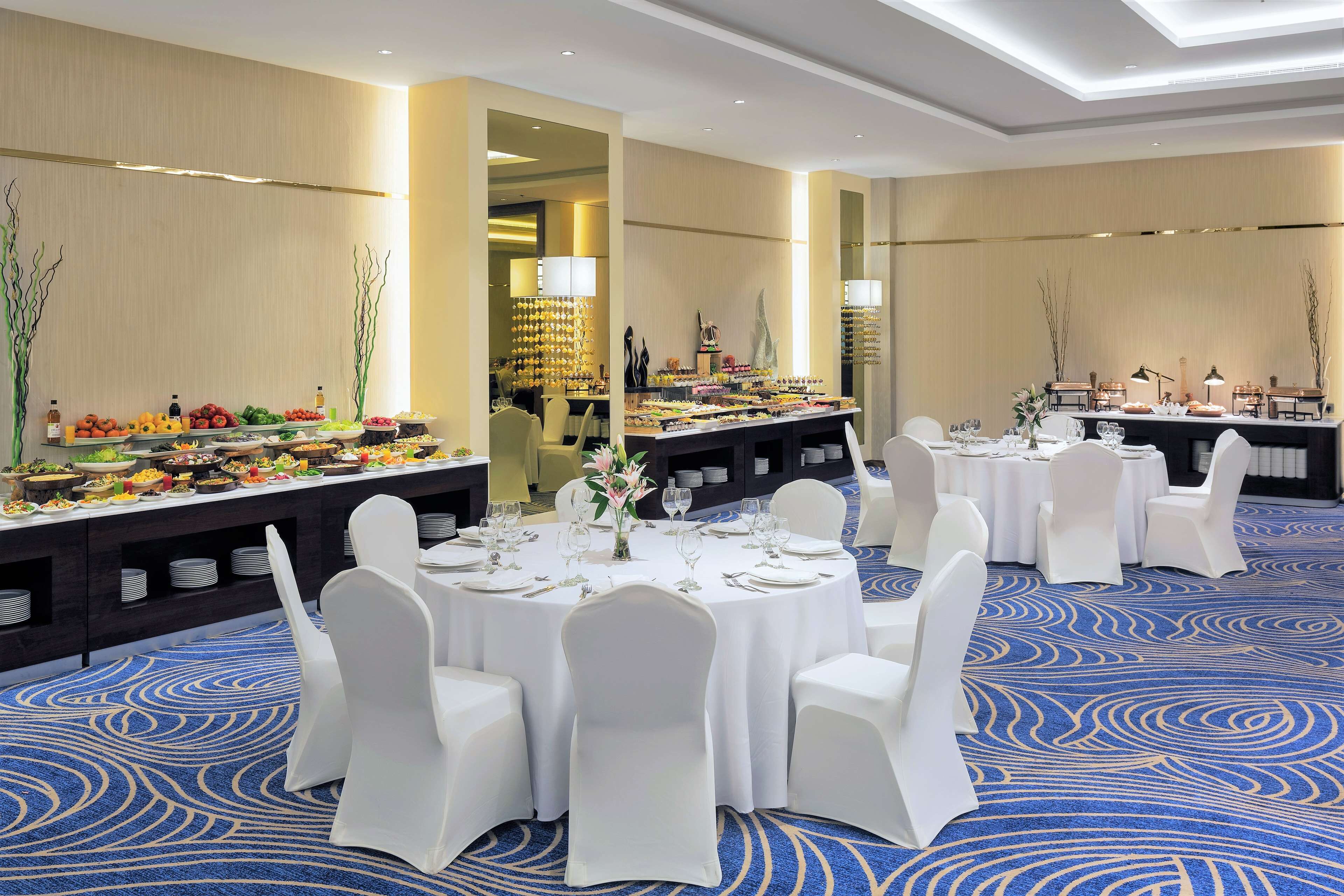 Un salón de banquetes con mesas redondas dispuestas para comer, sillas blancas y una zona de bufé con diversos alimentos.
