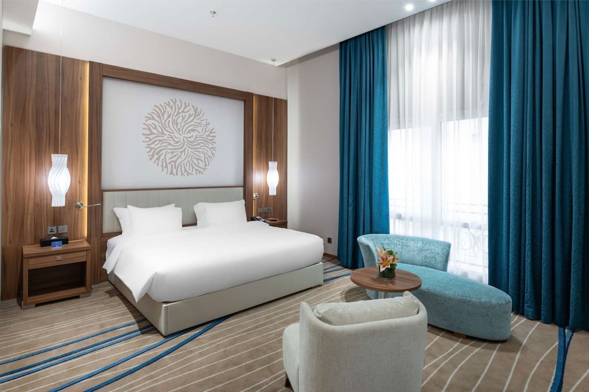 A hotel room with a large bed, a seating area with a chair and a small table, a decorative wall piece, and a window with curtains.