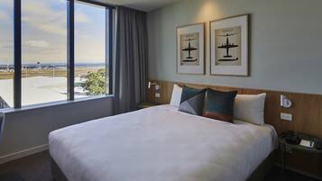 Een hotelkamer met een groot bed, uitzicht op het vliegveld en twee ingelijste foto's aan de muur.