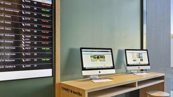 Un panneau d’affichage indiquant les vols et les numéros des portes d’embarquement, deux ordinateurs Apple iMac sur un bureau en bois, et une pancarte sur laquelle on peut lire : « CONNECT @ MANTRA ».