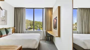 Quarto de hotel com duas camas, uma escrivaninha, uma cadeira, janela com cortinas e um quadro na parede.