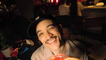 A person holding a cocktail glass with a red drink, seated at a table in a dimly lit indoor setting.