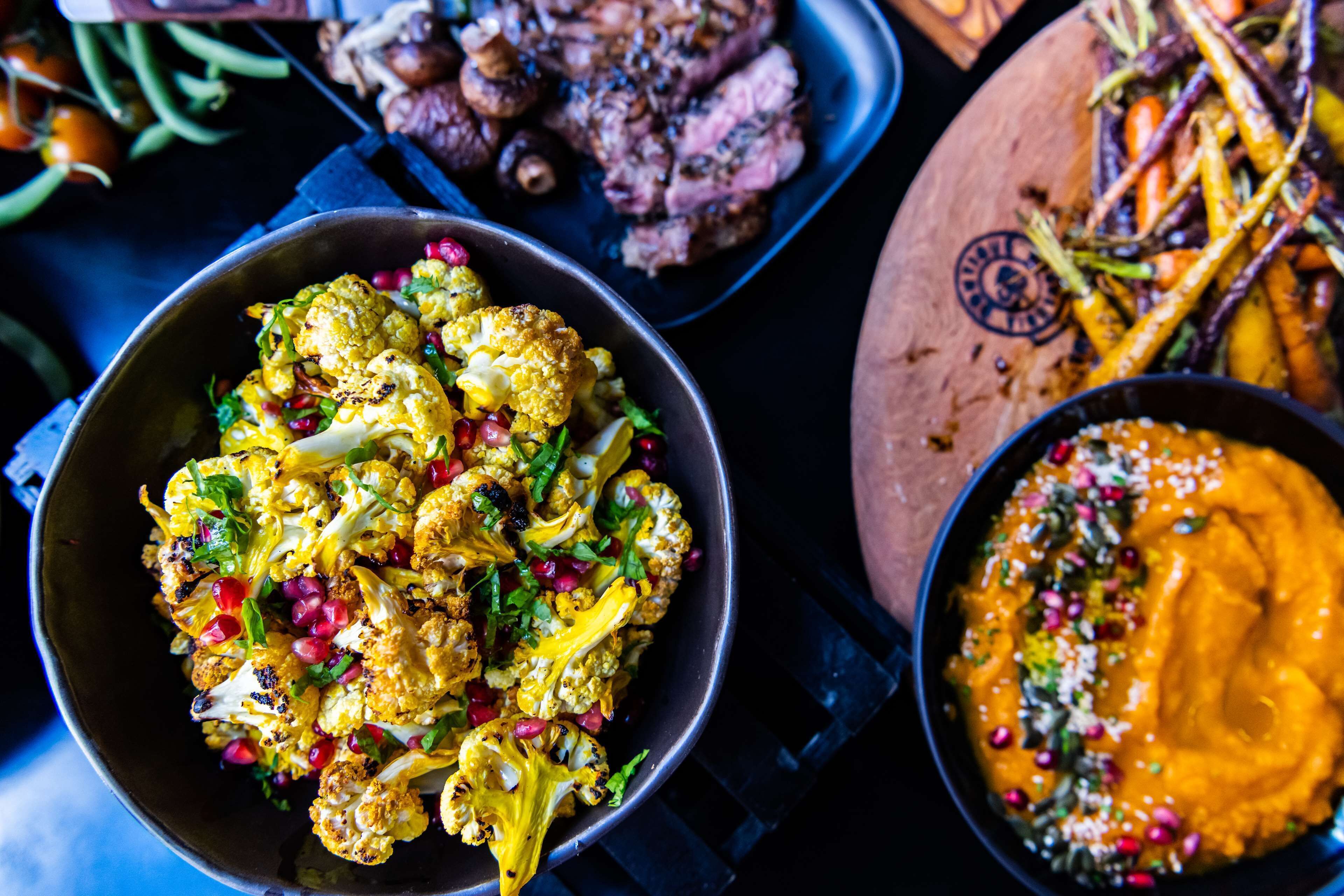 A variety of dishes including roasted cauliflower, a bowl of orange sauce, and a plate of grilled meat.