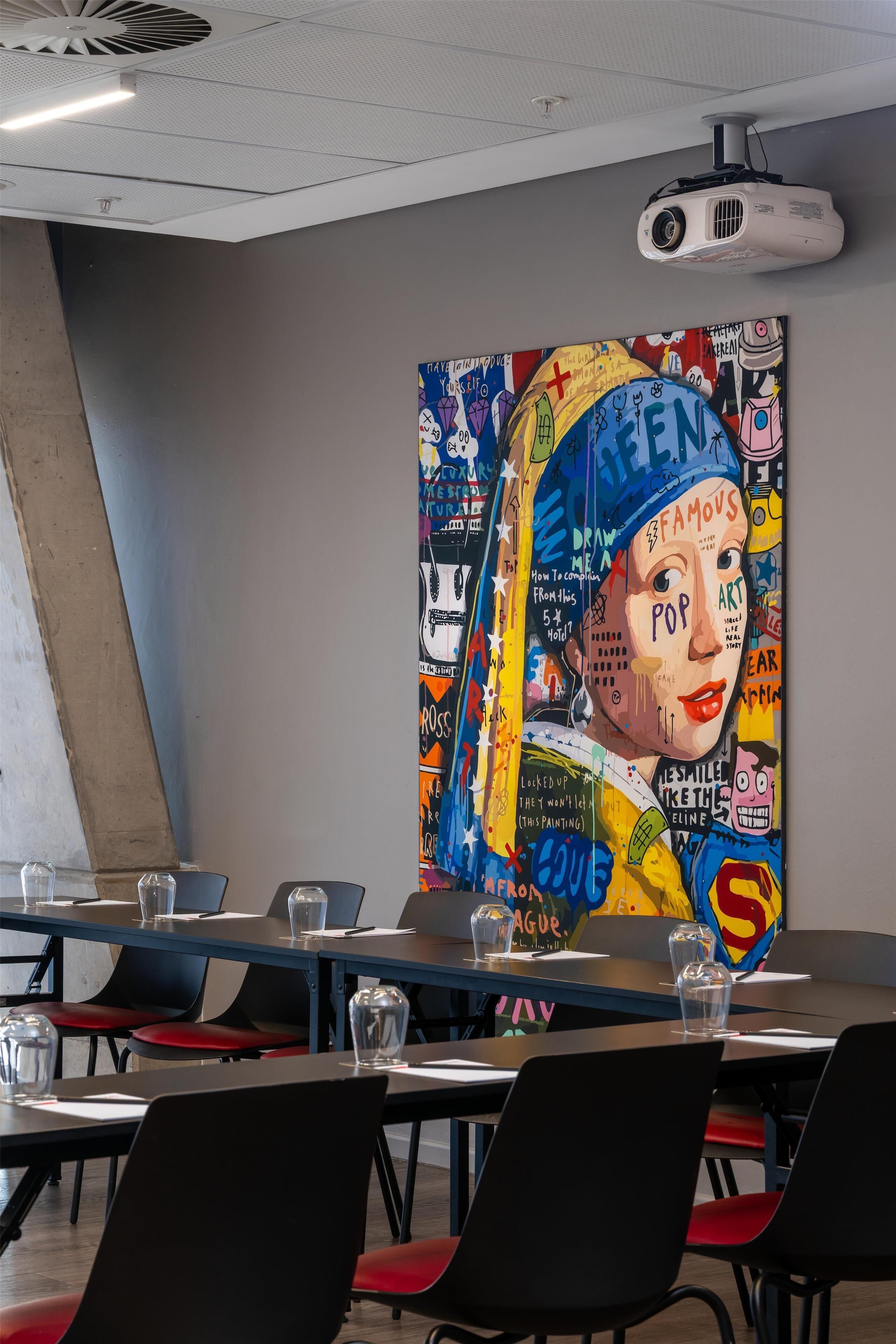 A conference room with a large colorful mural on the wall, a projector mounted on the ceiling, and a table with chairs.