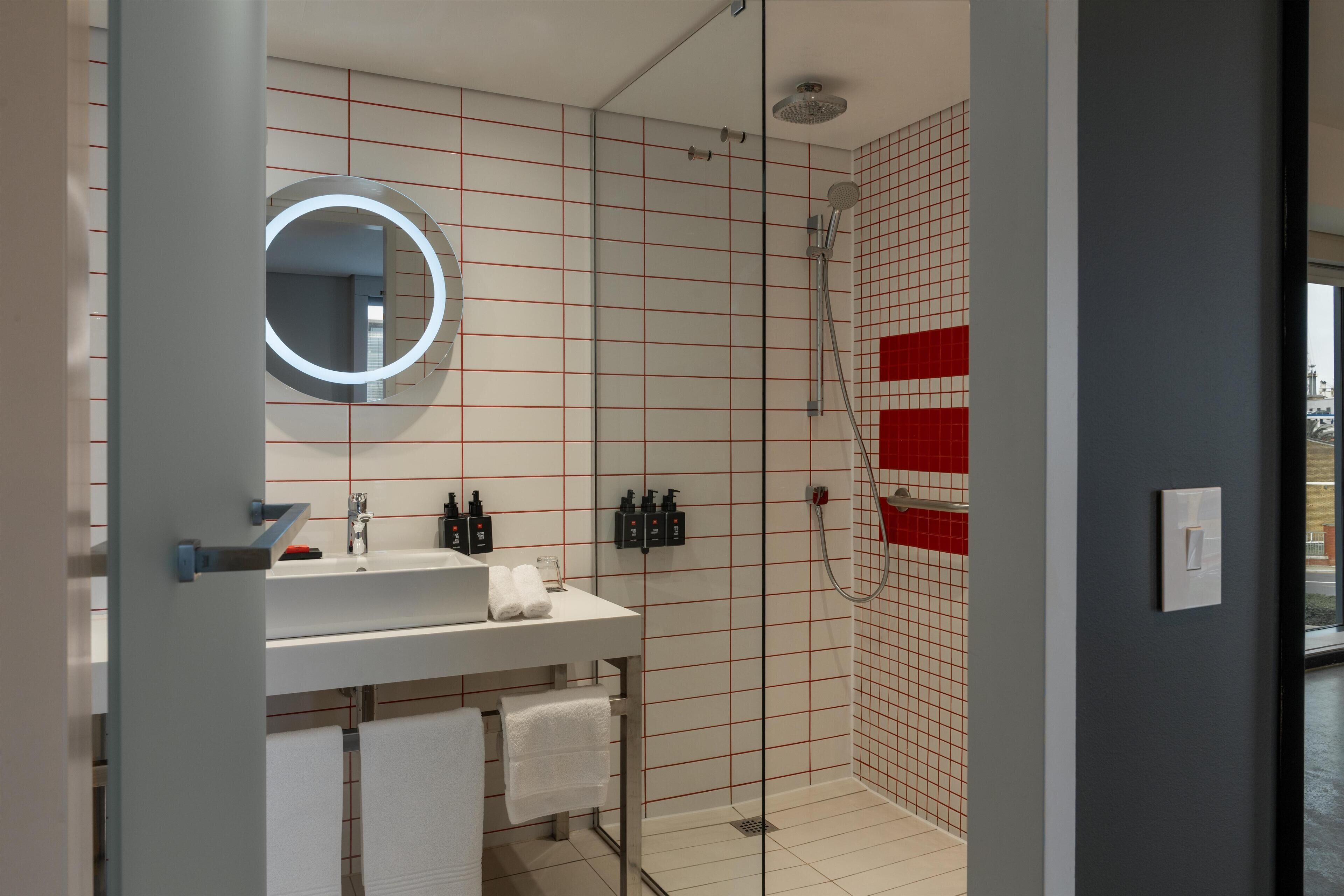 A modern bathroom with a glass shower enclosure, a white sink with a mirror, and red-tiled walls.