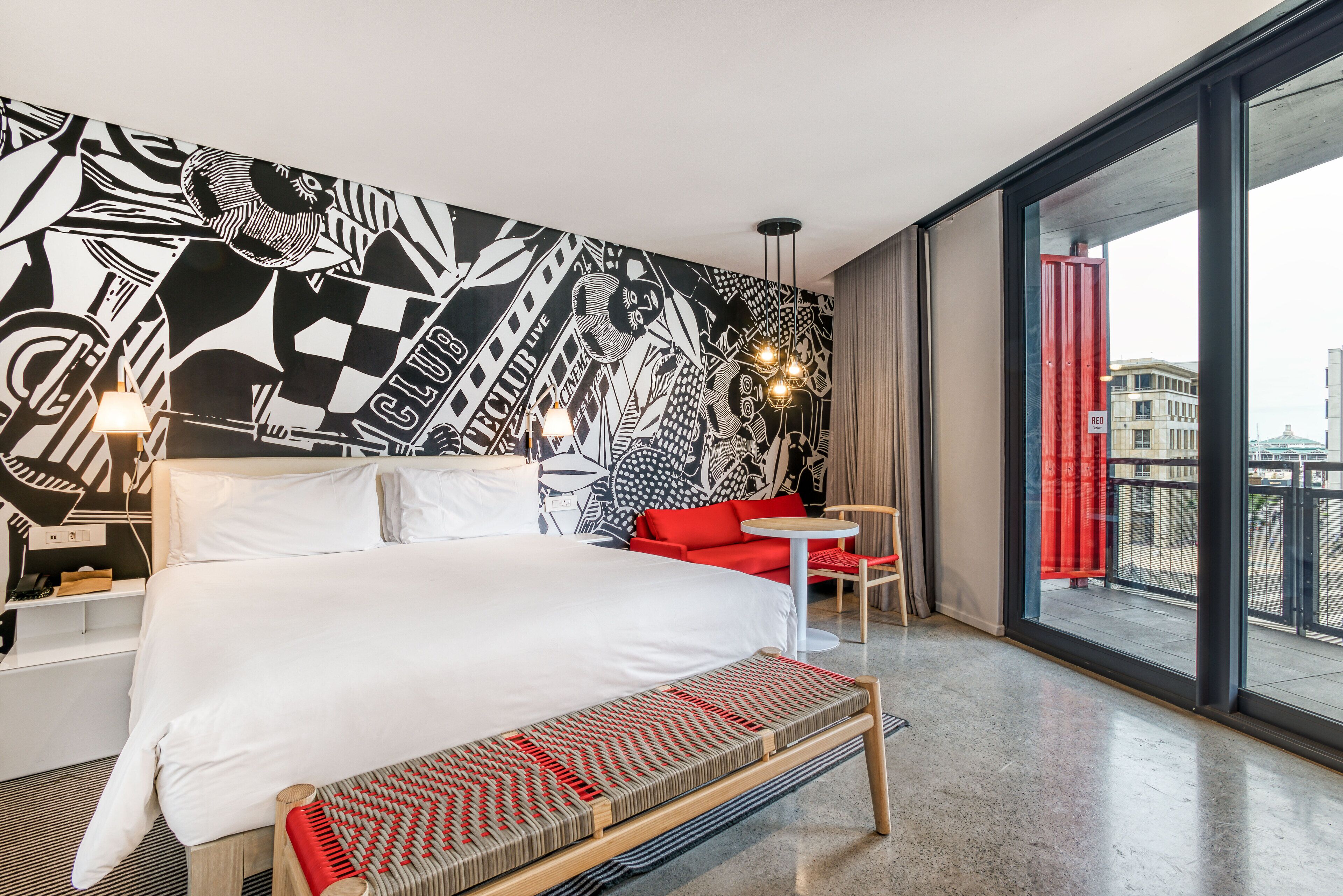 A modern hotel room with a large bed, a red sofa, a small table, and a large window with a view of the city.