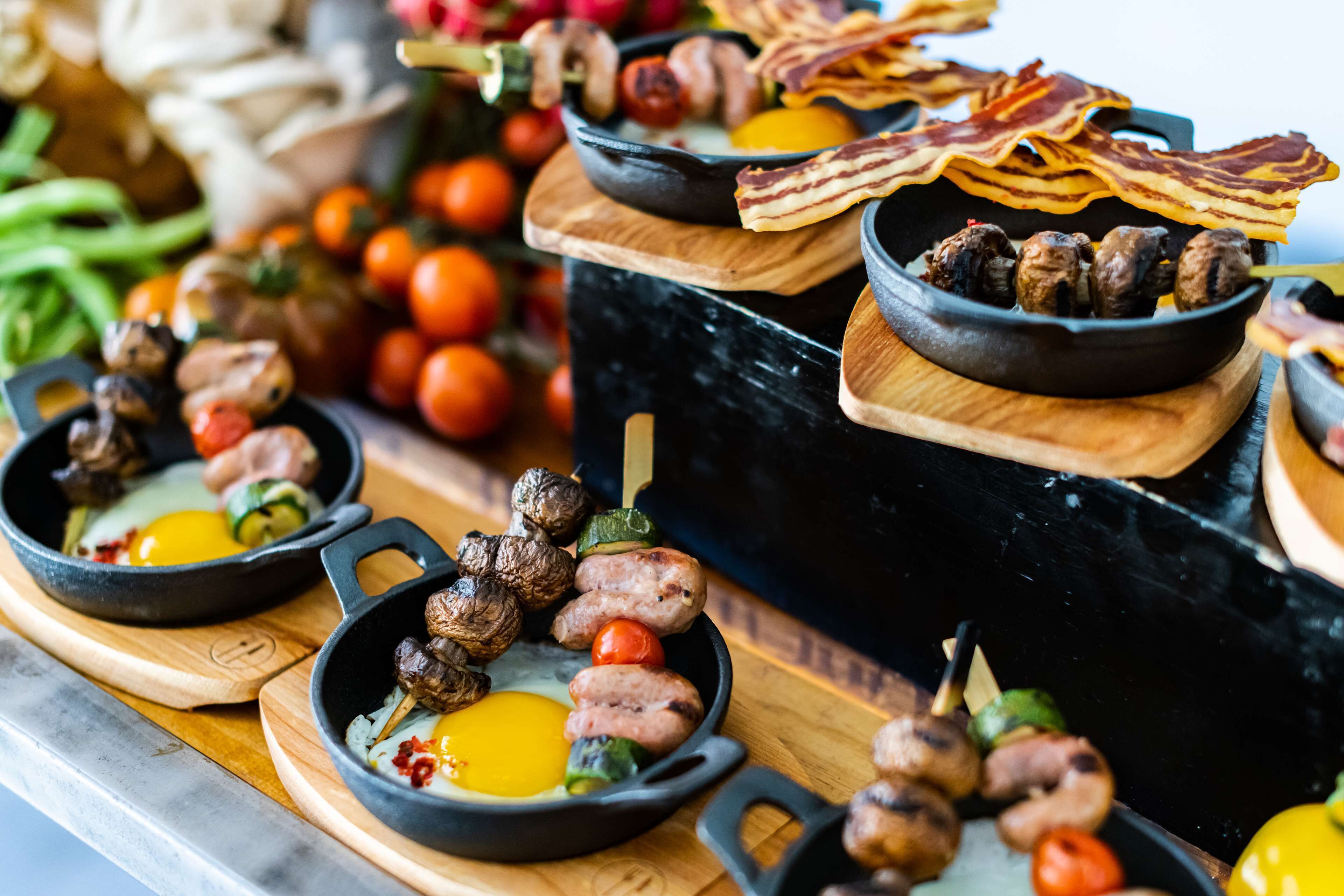 A variety of food items including sausages, eggs, and vegetables arranged on wooden boards and in cast iron pans.