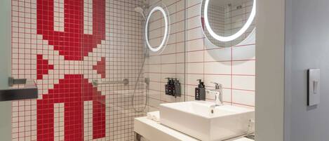 Une salle de bain moderne avec un mur recouvert de carreaux rouges et blancs disposés en motif quadrillé, un lavabo blanc avec un miroir au-dessus, et un espace douche doté d’un pommeau de douche à main.
