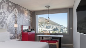 Una camera d'albergo con un letto, una scrivania, una sedia, un armadietto rosso, una finestra con vista sulla città e un muro decorato.