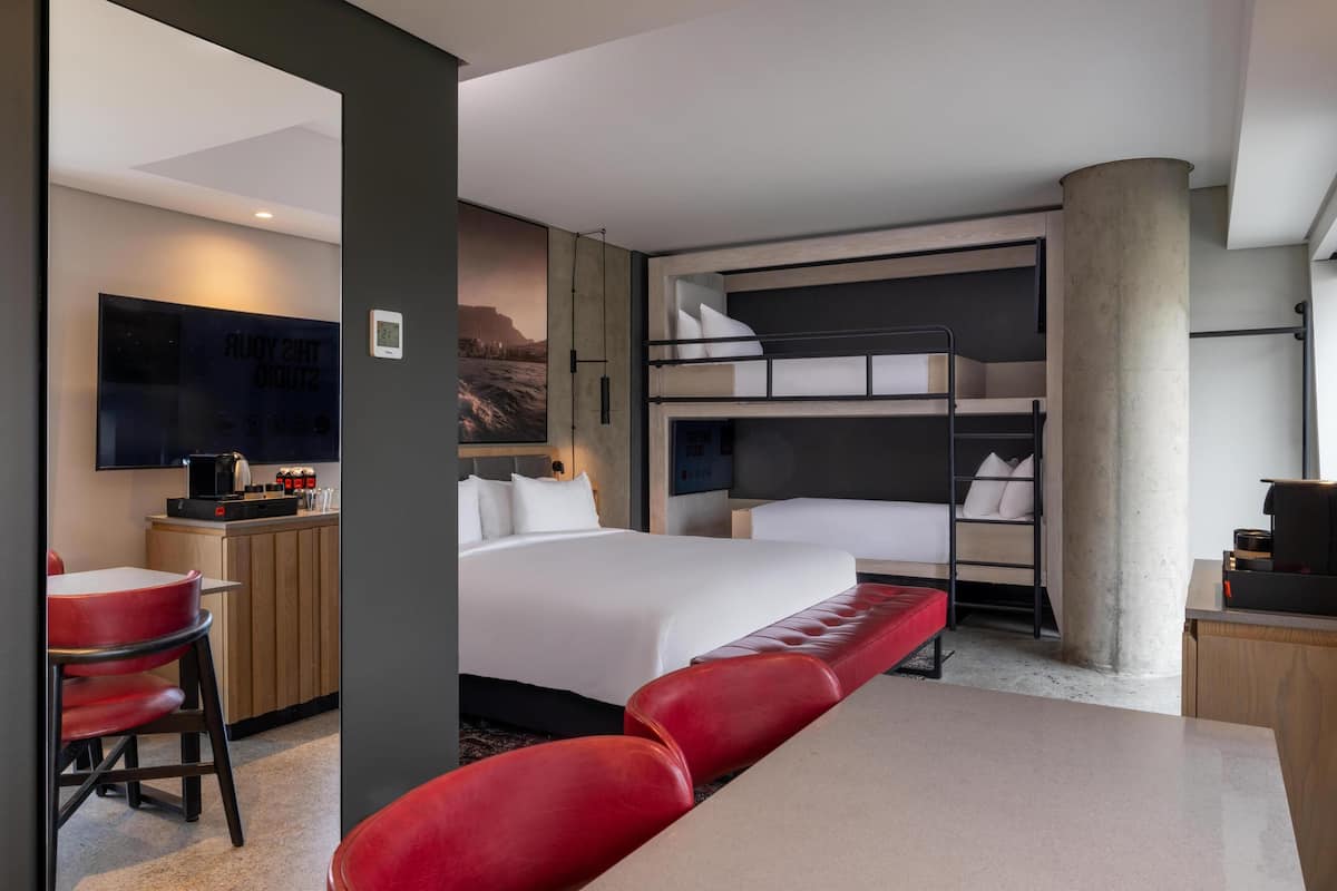 A modern hotel room with a large bed, a desk with a chair, a bunk bed, and a television.