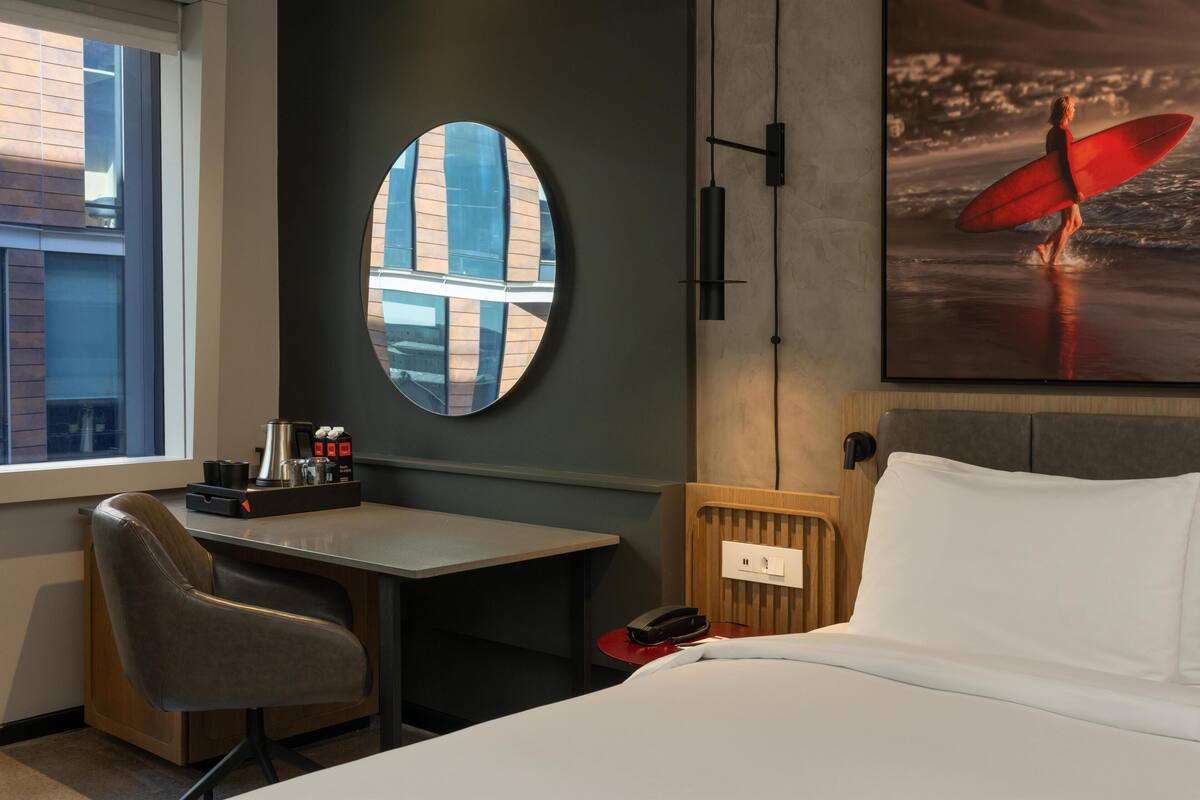 A hotel room with a bed, a desk, a chair, a round mirror, and a painting of a surfer on the beach.