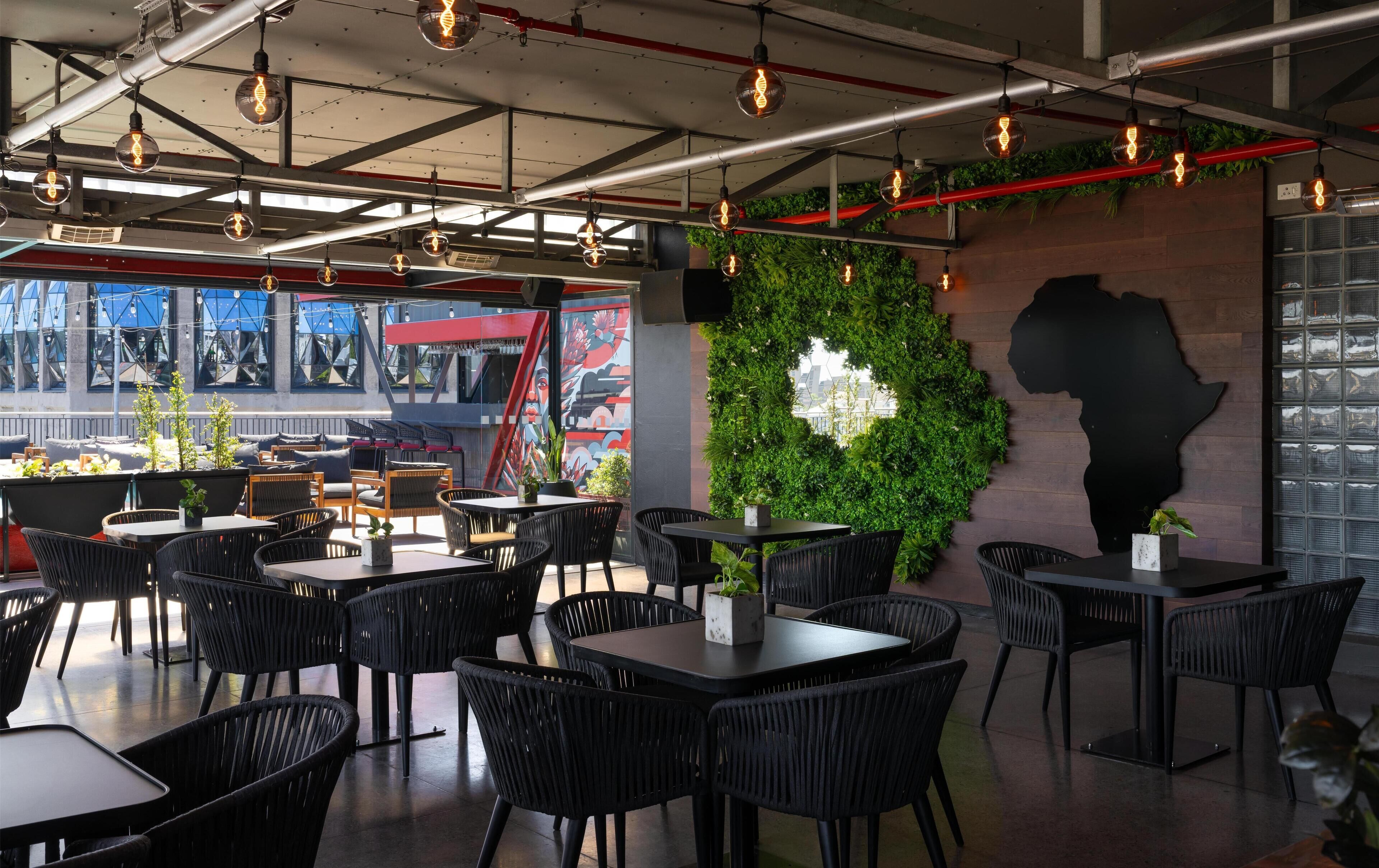 Een modern restaurant met zwarte rieten stoelen en tafels, een groene wand met een plant, en een muurschildering.
