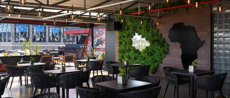 Un restaurante moderno con sillas y mesas de mimbre negro, una pared verde con una planta y un mural.