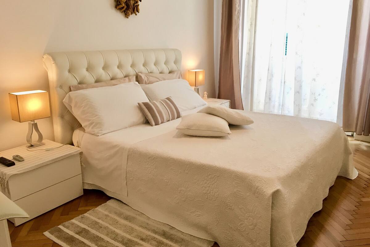 A neatly made bed with a tufted headboard, white bedding, and a striped area rug. There are two bedside tables with lamps and a decorative wall piece above the bed.