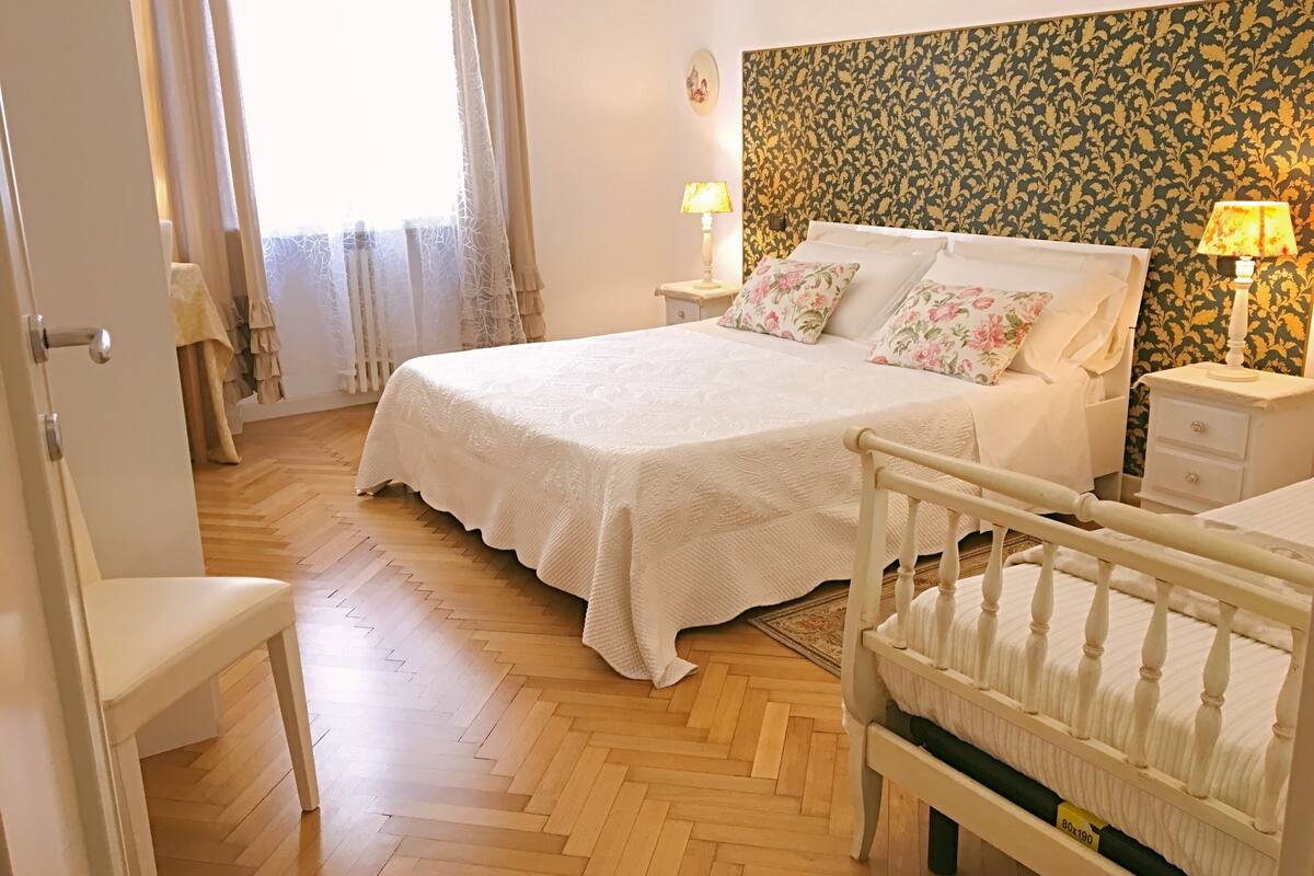 A bedroom with a large bed, two bedside tables with lamps, a wooden floor, and a patterned wallpaper on the wall.
