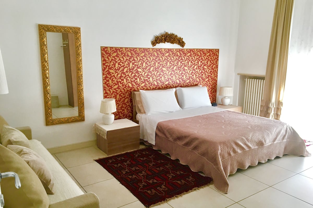 A hotel room with a bed, a nightstand, a lamp, a mirror, a red patterned headboard, a beige sofa, and a red patterned rug.