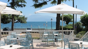 Un café al aire libre con mesas y sillas, sombrillas blancas y vista al mar.