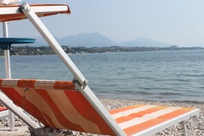 Una silla de playa con cojín a rayas, estructura metálica y cojín azul sobre un soporte, ubicada en una orilla pedregosa con un mar tranquilo y montañas distantes al fondo.