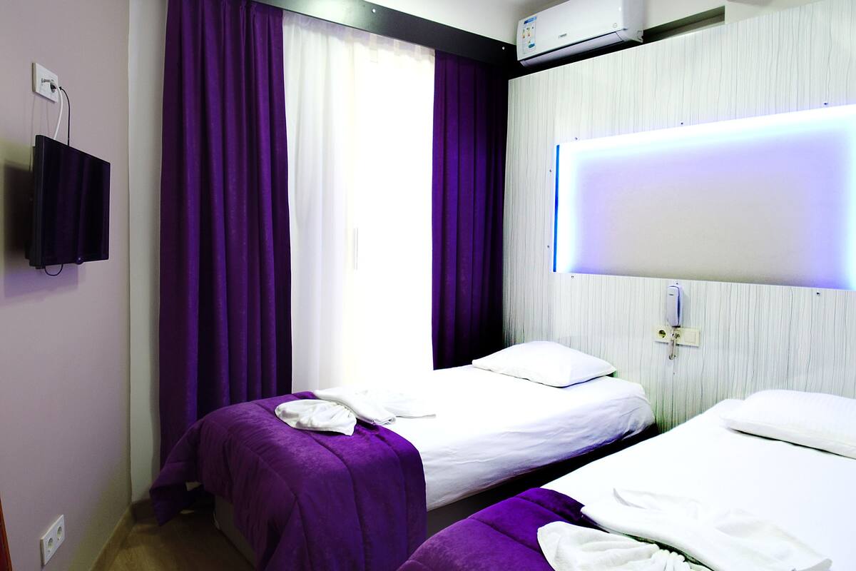 A hotel room with two beds, purple curtains, a mounted TV, and an air conditioning unit.