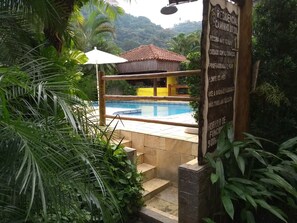 A pool area with a thatched-roof structure, surrounded by lush greenery and a signboard with text. - Residencial Caminho do Sol Chalés (São Sebastião)