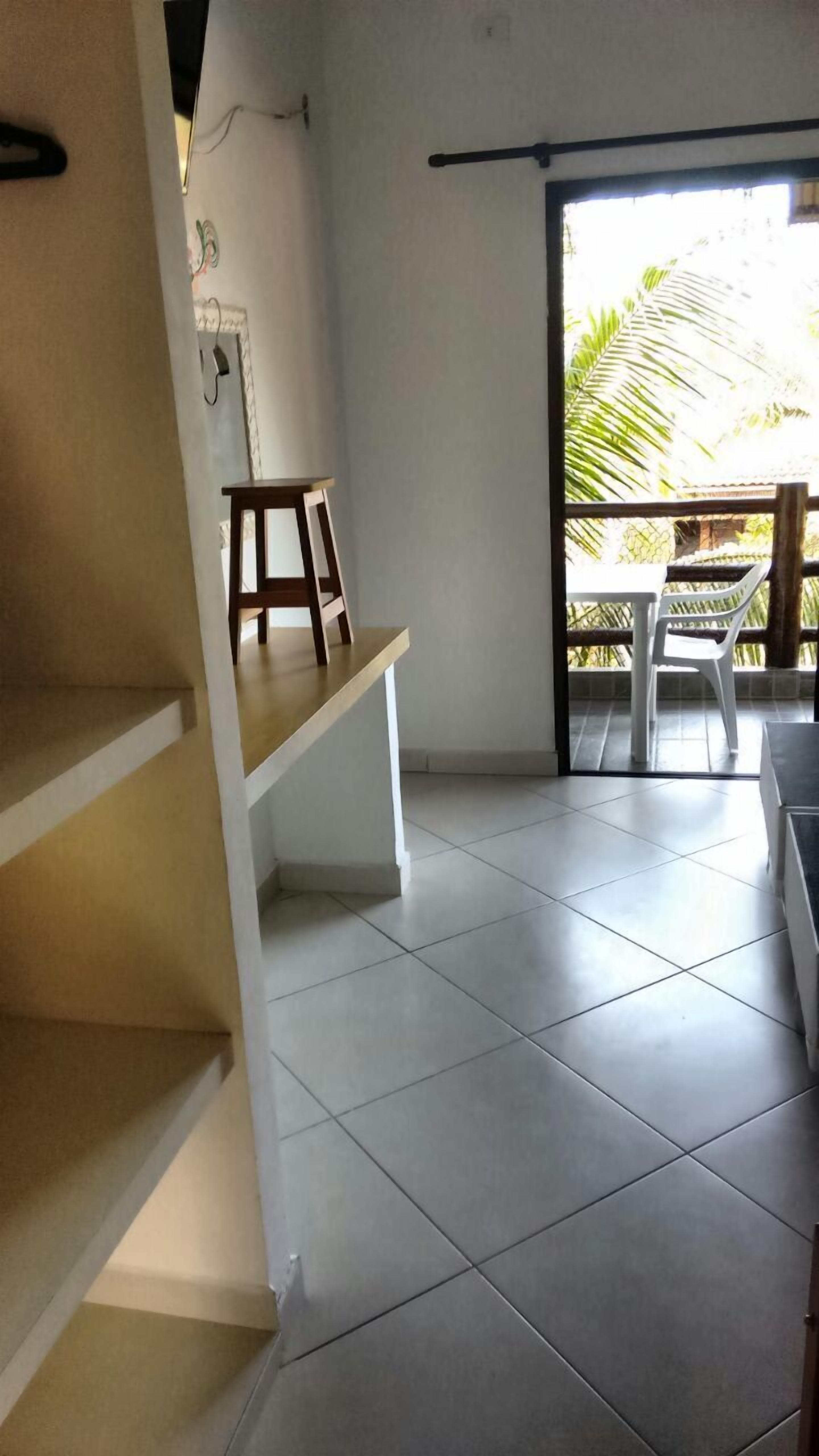A room with a tiled floor, a wooden bench, and a white chair near a sliding glass door leading to an outdoor area with a palm tree.