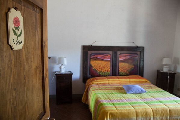 A bedroom with a bed, a nightstand, a wooden door, and a wall with two paintings. - Borgo Carpineto (Castelnuovo Berardenga)