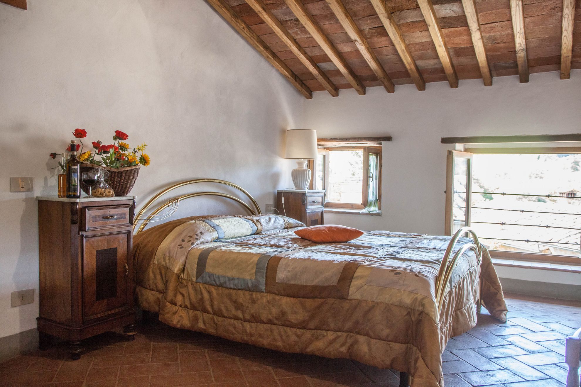A bedroom with a wooden ceiling, a bed with a quilt, a nightstand with a lamp, a window, and a wall-mounted shelf with flowers and bottles.