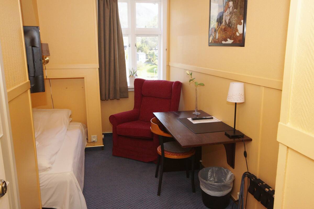 A compact hotel room with a single bed, a red armchair, a small table with a lamp, and a window with a view of greenery.
