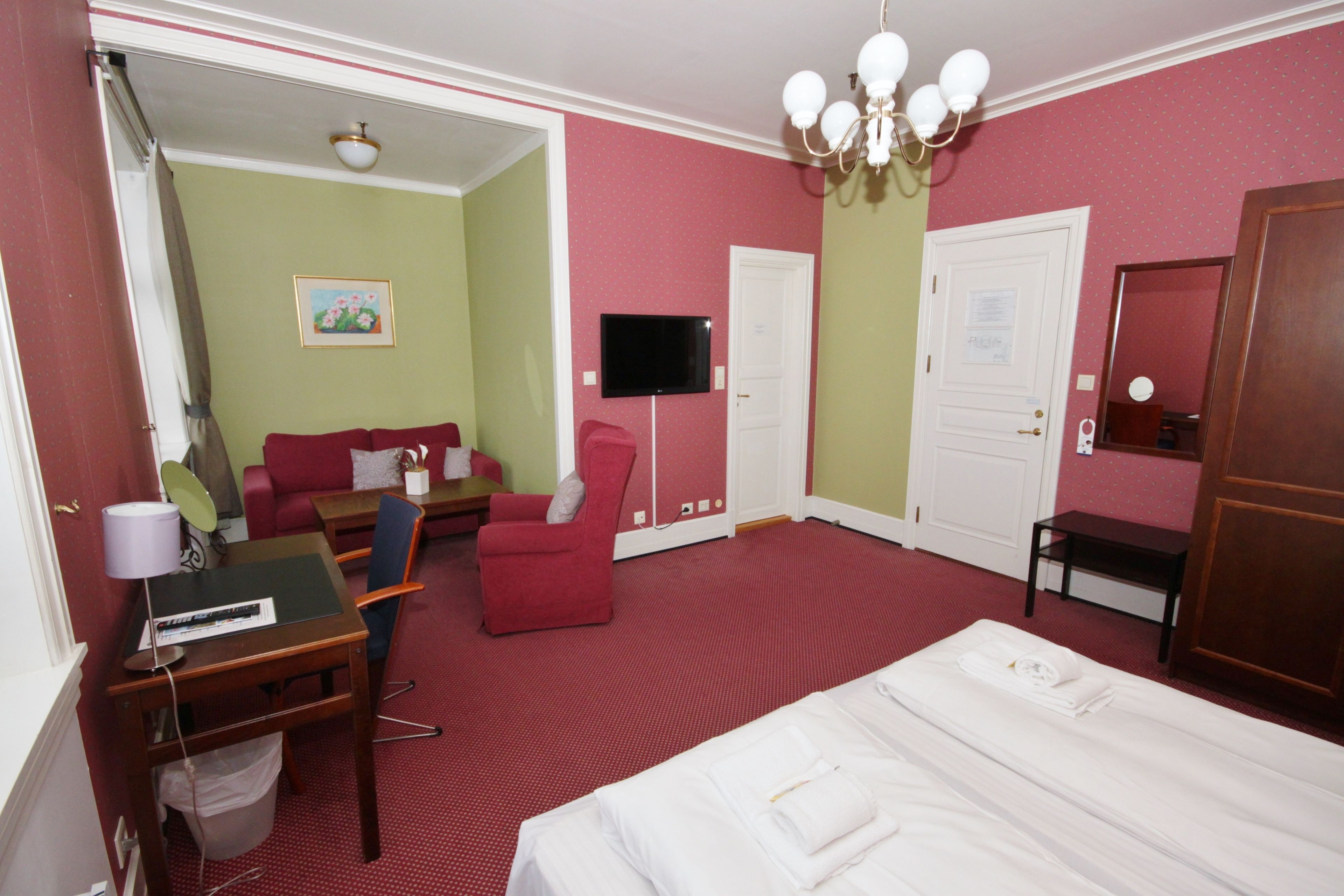 Una camera d'hotel con un letto, una scrivania con una sedia, una poltrona rossa, una televisione e una porta.