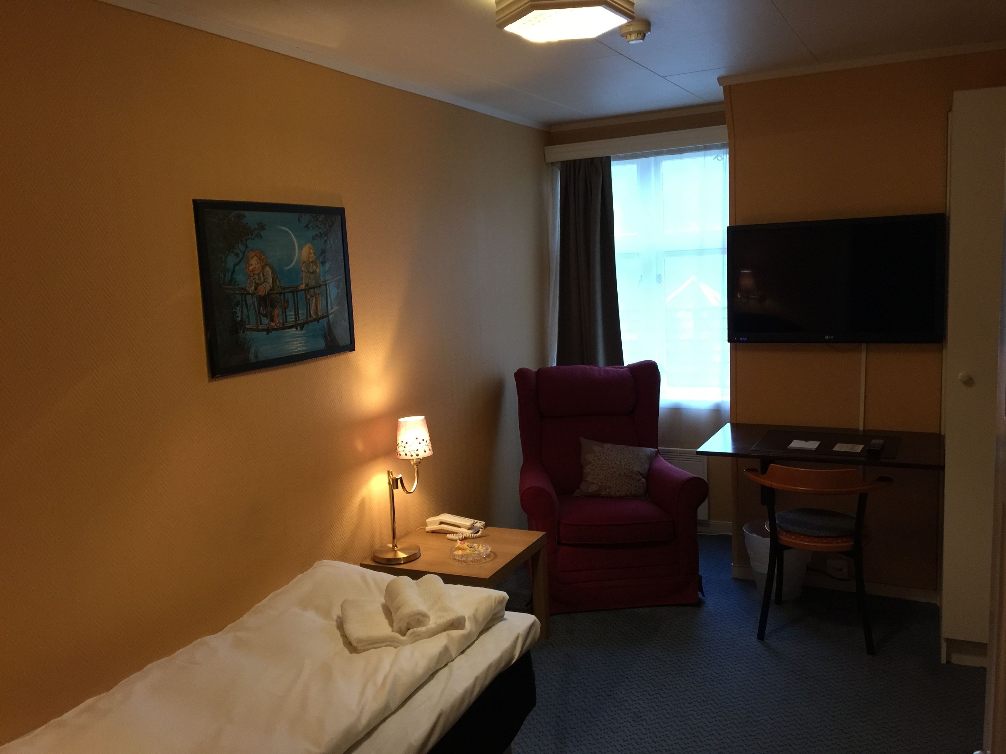 A hotel room with a bed, a red armchair, a desk with a chair, a TV, and a painting on the wall.