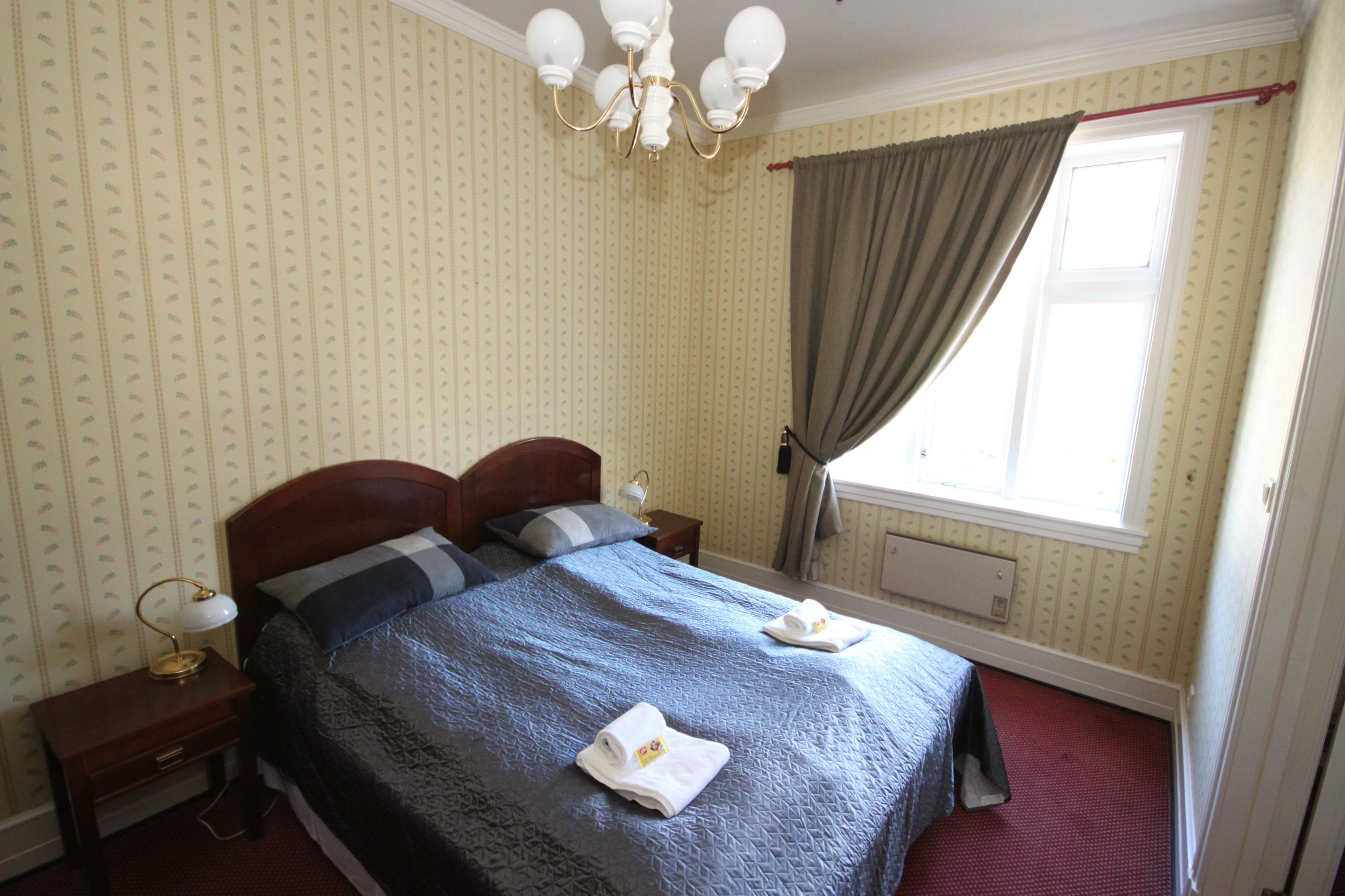 A neatly made bed with a blue bedspread and two pillows, a bedside lamp, and a window with curtains.