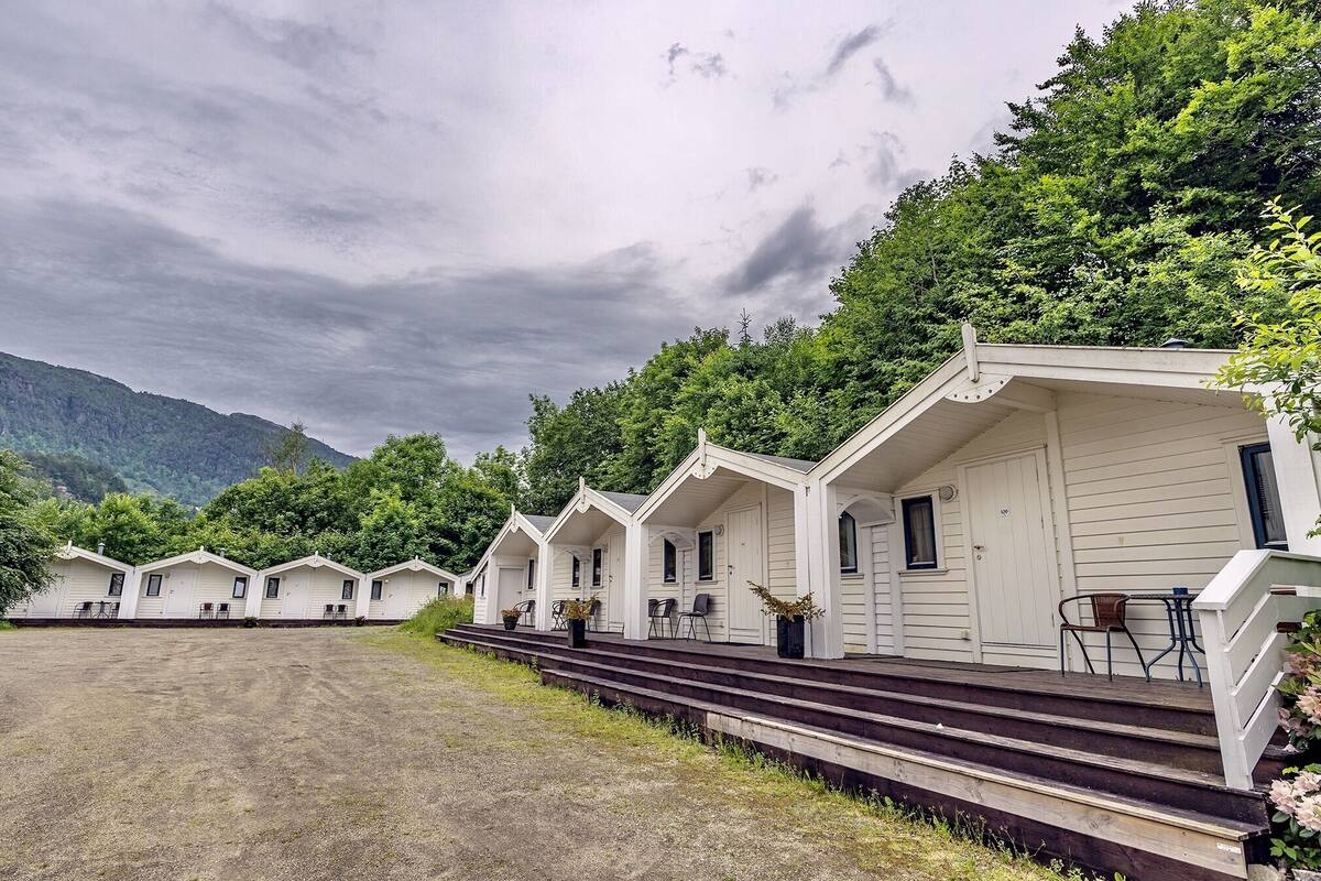 A row of white cabins with steps leading up to the entrance, surrounded by greenery and mountains in the background.