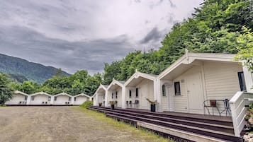 A row of white cabins with steps leading up to the entrance, surrounded by greenery and mountains in the background.